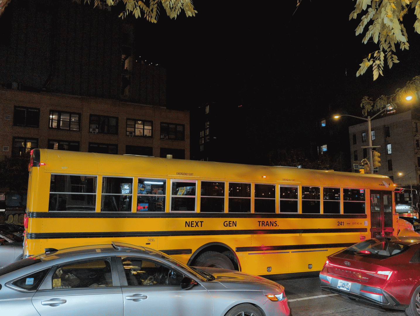 A US-ian schoolbus on a busy road between normal cars in a city. In bright yellow and instantly recognizablem the black letters on its side spell out NEXT GEN TRANS. on its side.